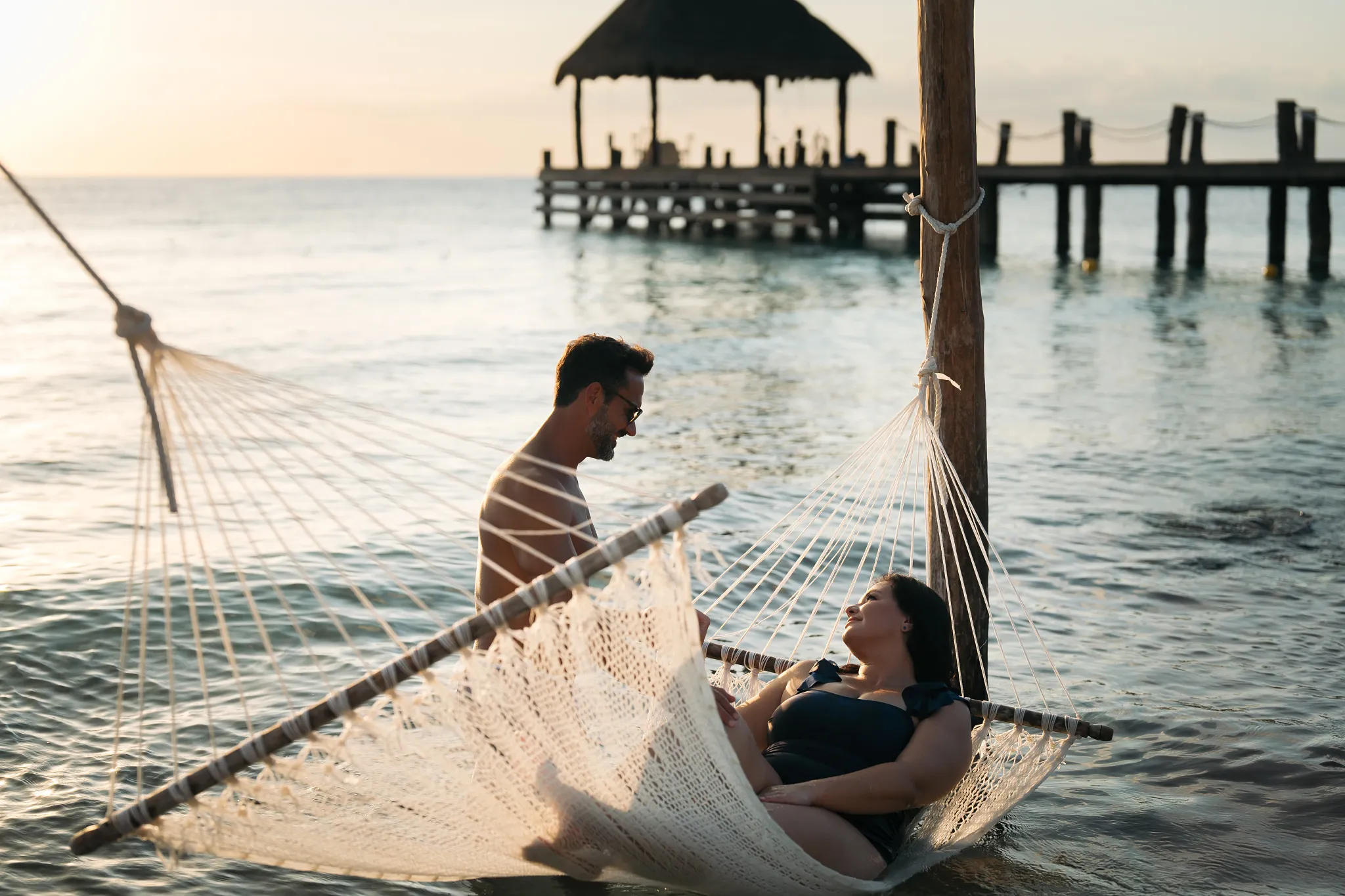 Couple relaxing together in a hammock over the water at Secrets Aura Cozumel during a golden sunset.