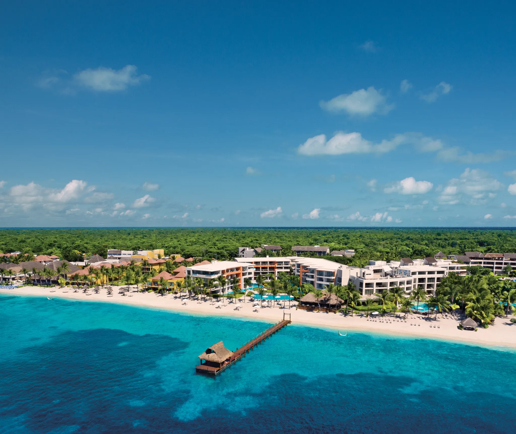 Panoramic aerial of Secrets Aura Cozumel with beachfront resort buildings, pier, and turquoise Caribbean waters.