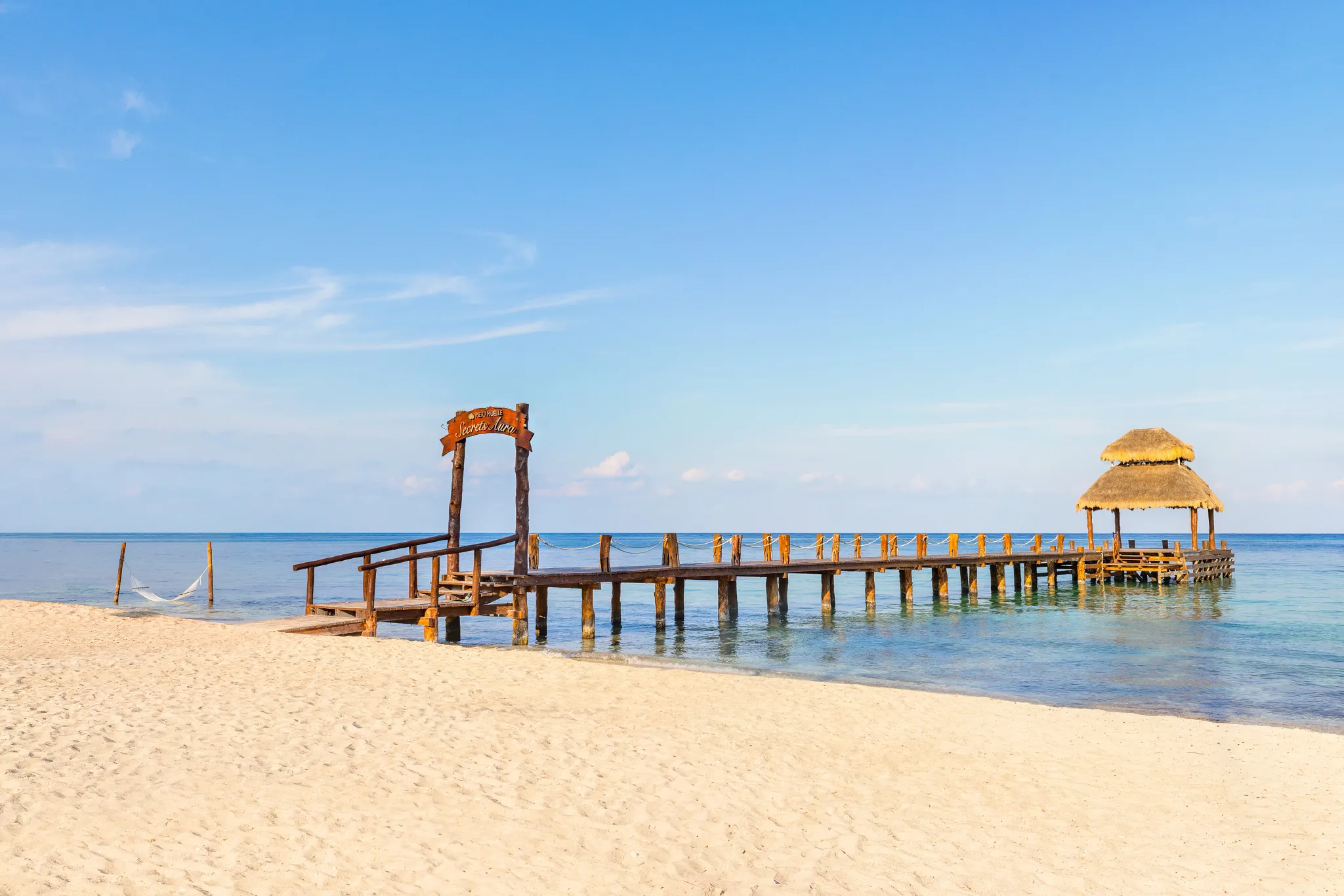 Wooden pier entrance on sandy beach at Secrets Aura Cozumel with turquoise Caribbean water.