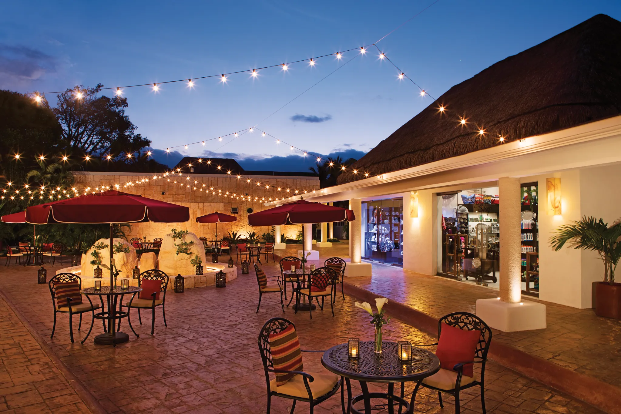 Outdoor plaza at Secrets Aura Cozumel at night with string lights, tables, and umbrellas.