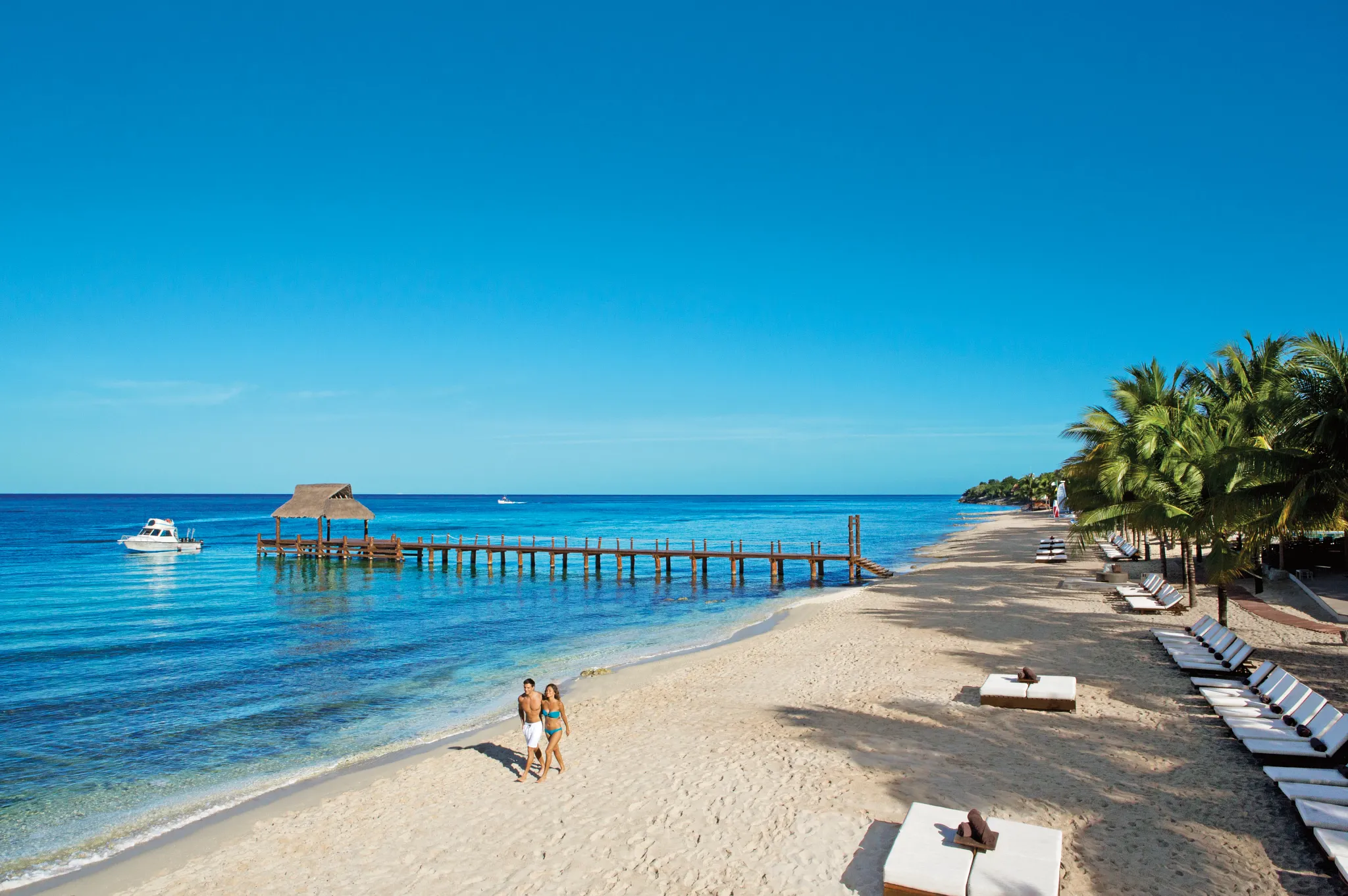Couple walking along sandy beach at Secrets Aura Cozumel with pier, lounge beds, and calm Caribbean waters.