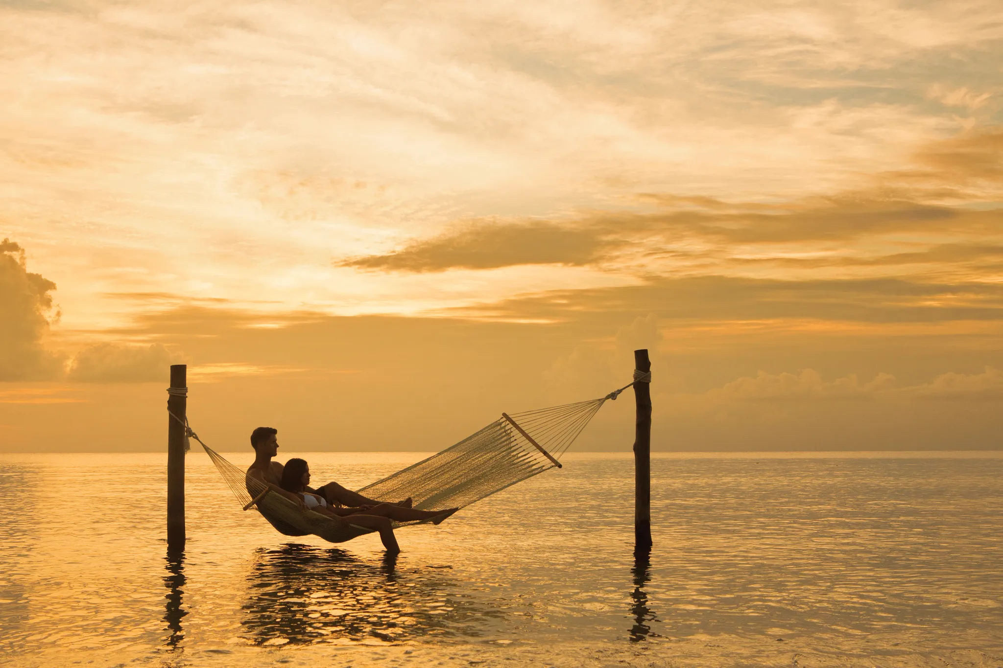Couple relaxing in hammock over shallow ocean water at sunset at Secrets Aura Cozumel.