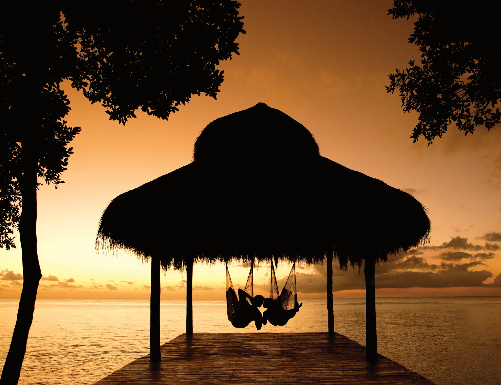 Couple relaxing in hammocks under a palapa at sunset over the ocean at Secrets Aura Cozumel, creating a romantic tropical scene.